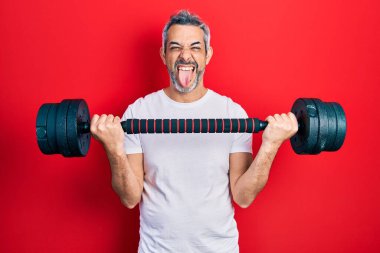 Handsome middle age man with grey hair wearing sportswear using dumbbells sticking tongue out happy with funny expression. 