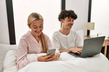 Dizüstü bilgisayar ve akıllı telefon kullanan genç ve güzel bir çift evdeki yatakta oturuyor..