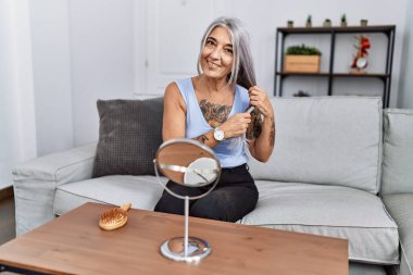 Orta yaşlı, kır saçlı, saçını tarayan, evdeki koltukta oturan bir kadın.