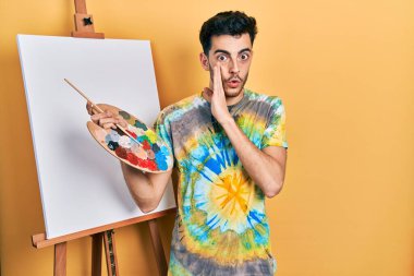 Young hispanic man standing drawing with palette by painter easel stand hand on mouth telling secret rumor, whispering malicious talk conversation 