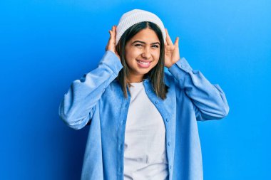 Şirin yün şapkalı genç Latin kadın iki elini de kulak hareketlerinde duymaya çalışıyor, dedikoduya meraklı. İşitme sorunu, sağır 