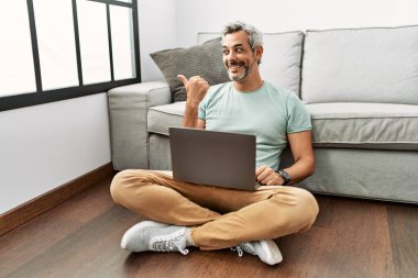 Middle age hispanic man using laptop sitting on the floor at the living room smiling with happy face looking and pointing to the side with thumb up. 