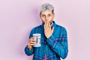 Modern boyalı saçları olan genç İspanyol bir adam domuz kumbarasını elinde tutuyor ağzını eliyle kapatıyor, şaşırmış ve hata yapmaktan korkuyor. şaşırmış ifade 