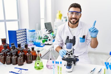 Handsome hispanic man working at scientist laboratory smiling happy pointing with hand and finger to the side 