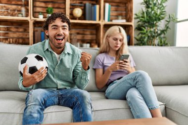 Adam mutlu bir şekilde futbol maçı izliyor ve kız arkadaşı evde akıllı telefon kullanıyor..