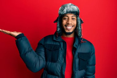 Sakallı, kışlık şapkalı, ceketli, neşeli gülümseyen ve avuç içi kameraya bakan Afro-Amerikan genç adam.. 