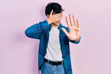 Yakışıklı hippi genç adam gündelik beyaz tişört ve klasik gömlek giyip gözlerini ellerle kapatıyor ve üzgün ve korku ifadeleriyle jest yapıyor. utangaç ve olumsuz kavram. 