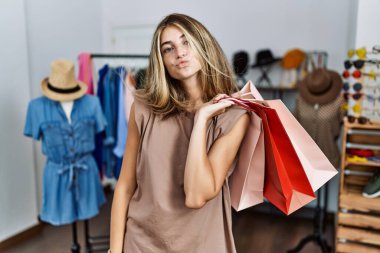 Young blonde woman holding shopping bags at retail shop looking at the camera blowing a kiss on air being lovely and sexy. love expression. 