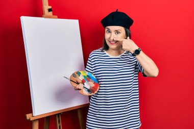 Orta yaşlı İspanyol kadın paletle resim çiziyor, resim sehpası ayakta el parmakları ve burunla işaret ediyor, neşeyle gülümsüyor. güzellik konsepti 