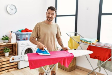 Genç İspanyol adam, çamaşırhanede ütü yaparken gülümsüyor.