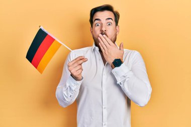 Handsome caucasian man with beard holding germany flag covering mouth with hand, shocked and afraid for mistake. surprised expression 