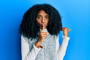 Afro saçlı, kışlık süveter giyen Afro-Amerikalı bir kadın sessiz olmak istiyor. Parmakları dudaklarında ve elleri yanda. sessizlik ve gizli konsept. 