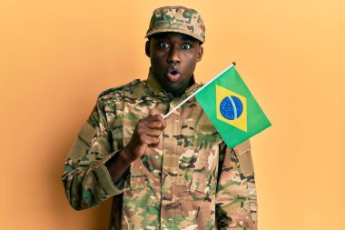 Asker üniforması giyen genç Afro-Amerikan bir adam elinde Brezilya bayrağı tutarken ağzı açık kalmış, korkmuş ve şaşkın bir şekilde şaşırmış. 