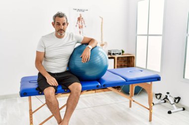 Middle age hispanic man at pain recovery clinic holding pilates ball looking sleepy and tired, exhausted for fatigue and hangover, lazy eyes in the morning. 
