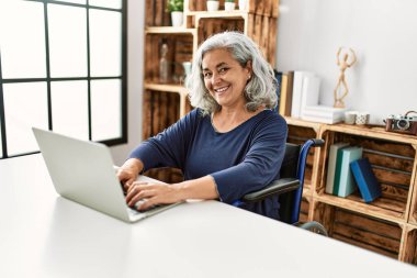 Evde tekerlekli sandalyede oturan orta yaşlı, gri saçlı, engelli bir kadın..