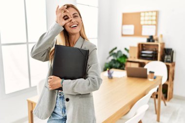 Blonde business woman at the office doing ok gesture with hand smiling, eye looking through fingers with happy face. 