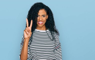 Orta yaşlı Afro-Amerikalı kadın gündelik giysiler giyip iki numarayı gösterirken kendine güvenen ve mutlu bir şekilde gülümsüyor.. 