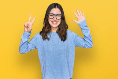 Genç ve güzel bir kadın günlük kıyafetler ve gözlük takıyor. Yedi numaralı parmaklarıyla işaret ediyor ve gülümsüyor. Kendinden emin ve mutlu.. 