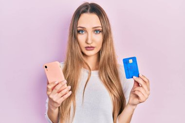 Young blonde girl holding smartphone and credit card relaxed with serious expression on face. simple and natural looking at the camera. 