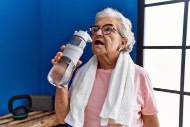 Spor kıyafetleri giymiş, spor salonunda elinde su şişesi tutan yaşlı, kır saçlı bir kadın.