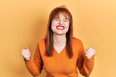 Kızıl saçlı, turuncu süveter giyen çok mutlu ve heyecanlı kolları havada, gülümseyen ve başarı için bağıran bir jest yapan genç kadın. kutlama konsepti. 