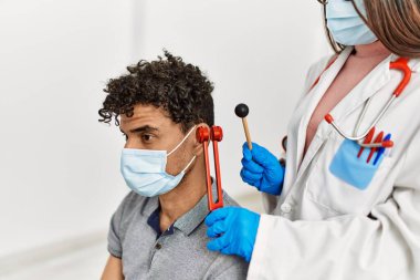 Tıbbi maske takan genç latin doktor kadın muayene odasında diapason kullanan bir adamın kulağını kesiyor..