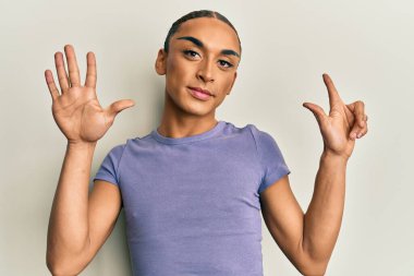 Makyaj yapan, uzun saçlı, günlük tişörtlü, 7 numaralı parmaklarıyla gösteriş yapan, kendinden emin ve mutlu bir şekilde gülümseyen İspanyol.. 