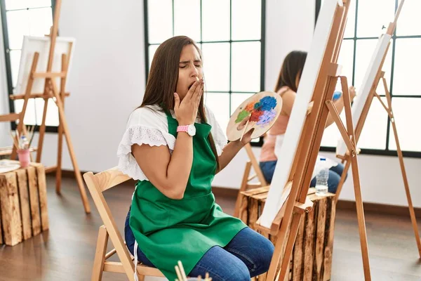 若いヒスパニック系のアーティストの女性がアートスタジオでキャンバスに絵を描く手で覆われた口の疲れあくび退屈 落ち着きもなく眠気もなく — ストック写真