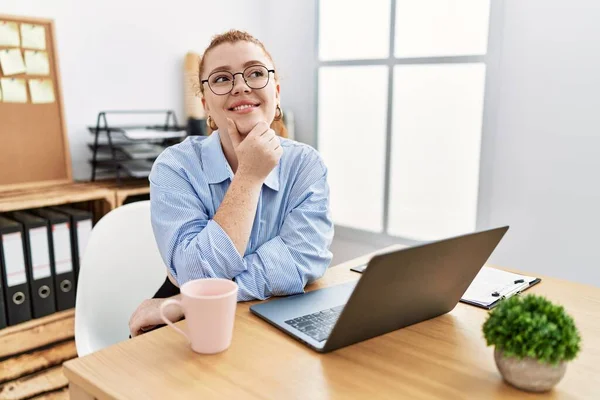 年轻的红头发女人在办公室里用电脑笔记本电脑 手托着下巴思考问题 沉思的表情 微笑和体贴的脸 怀疑概念 — 图库照片