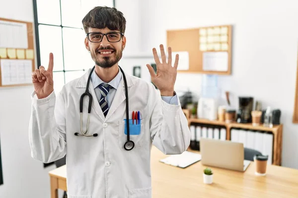 在办公室里 留着胡子 身穿医生制服和听诊器的西班牙裔男子露出并用手指指点着6号 同时带着自信和快乐的笑容 — 图库照片
