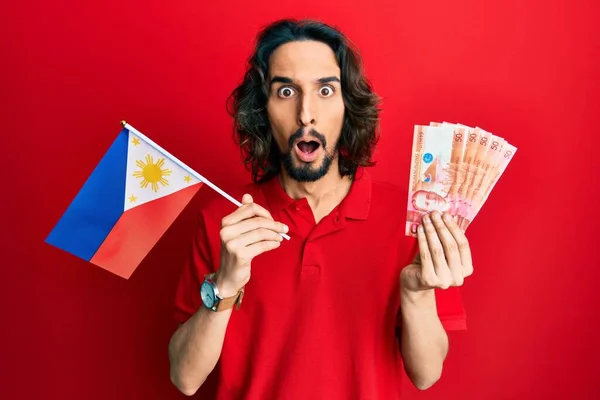 Young Hispanic Man Holding Philippines Flag Pesos Banknotes Afraid Shocked — Stock Photo, Image