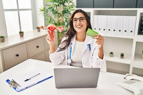 Young hispanic doctor woman holding anatomical female genital organ and birth control pills smiling and laughing hard out loud because funny crazy joke.