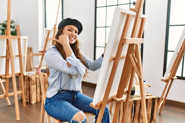 若いヒスパニック系のアーティストの女性は 手と親指で後ろを指しているアートスタジオでキャンバスに絵を描いて 自信を持って笑って — ストック写真