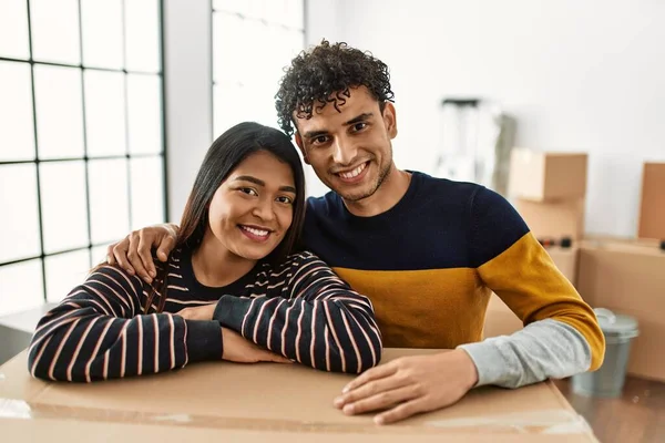 Genç Latin çift mutlu bir şekilde gülümsüyor. Yeni evlerinde karton kutuya yaslanıyorlar..
