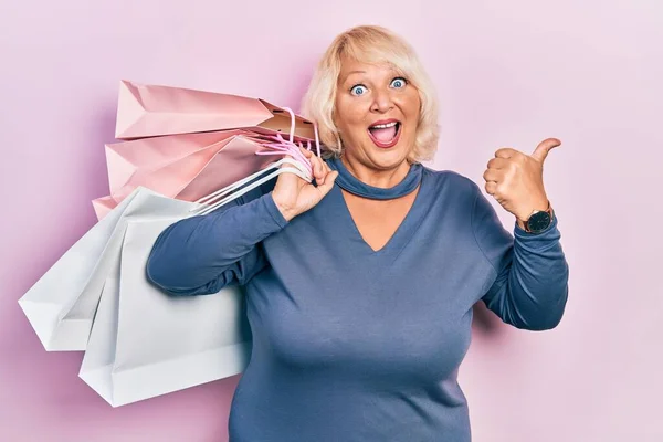 Medelålders Blond Kvinna Håller Shoppingpåsar Pekar Tummen Upp Till Sidan — Stockfoto