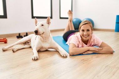 Beyazlı genç bir kadın spor merkezinde yoga minderine uzanmış kendinden emin bir şekilde gülümsüyor.