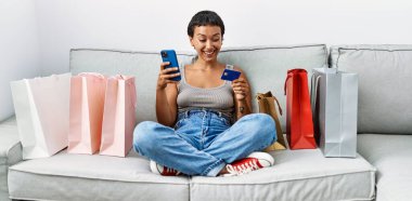 Young hispanic woman using smartphone and credit card shopping at home clipart