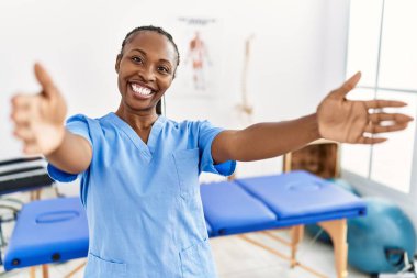 Saçları örgülü siyah bir kadın, ağrı kesici kliniğinde çalışıyor kameraya bakıyor ve sarılmak için kollarını açmış gülümsüyor. Mutluluğu kucaklayan neşeli bir ifade.. 