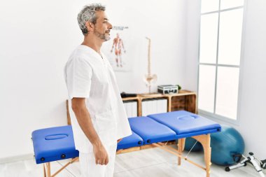 Middle age hispanic therapist man working at pain recovery clinic looking to side, relax profile pose with natural face and confident smile. 
