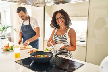Orta yaşlı İspanyol çift, gülümsüyor. Mutfakta tavaya yiyecek koyuyorlar.