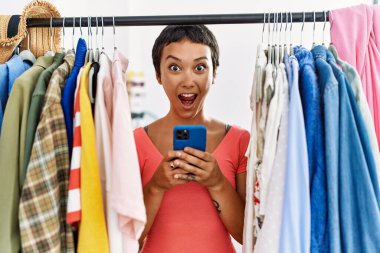 Kısa saçlı genç İspanyol kadın elbise rafında kıyafet arıyor. Akıllı telefonu kullanıyor. Şaşırmak için ağzı açık ve korkmuş. 