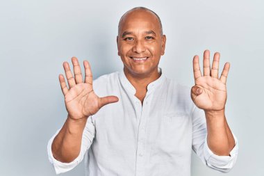 Orta yaşlı, gündelik kıyafetler giyen Latin bir adam dokuz numarayı gösterip işaret ederken kendinden emin ve mutlu bir şekilde gülümsüyor.. 