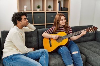 Mutlu gülümseyen genç çift evdeki kanepede oturup klasik gitar çalıyor..