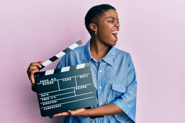 Young african american woman holding video film clapboard angry and mad screaming frustrated and furious, shouting with anger. rage and aggressive concept. 