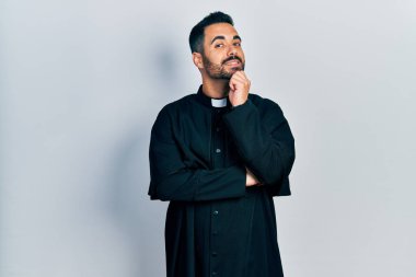 Sakallı, Katolik rahip cübbesi giyen yakışıklı, kameraya kendinden emin, kolları çaprazlanmış ve elleri çenesinde gülümseyen bir adam. Olumlu düşünmek. 