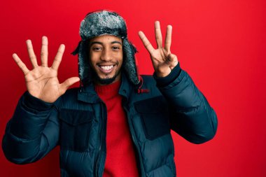 Sakallı, kışlık şapkalı ve paltolu genç Afrikalı Amerikalı sekiz numaralı parmaklarıyla işaret ederken kendinden emin ve mutlu bir şekilde gülümsüyor.. 