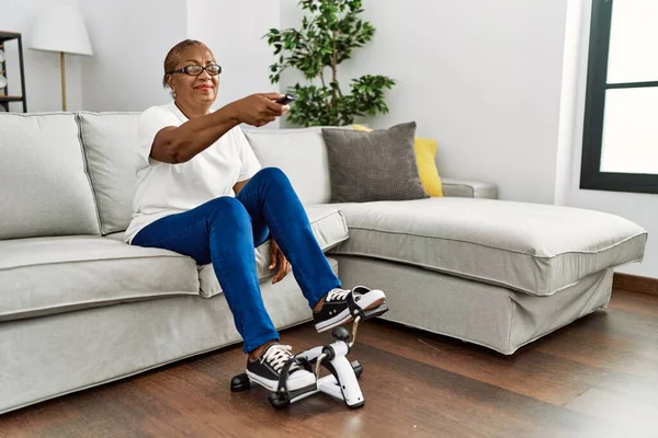 Senior african american woman watching tv doing sport using bike pedals machine at home