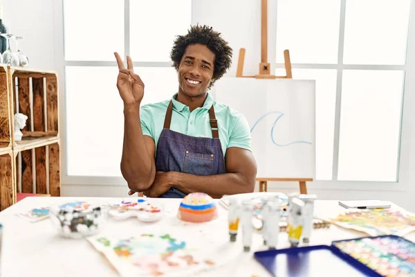若いアフリカ系アメリカ人の男性がアートスタジオのテーブルの上に座り カメラに向かって勝利の合図をして 幸せそうな顔をして微笑んだ — ストック写真