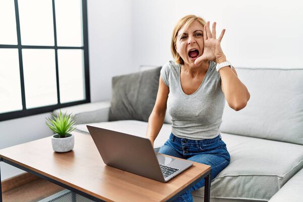 Блондинка средних лет использует ноутбук дома кричать и громко кричать в сторону с рукой в рот. коммуникационная концепция. 