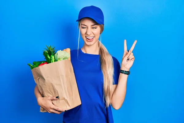 年轻的高加索女人穿着信使制服 带着食品杂货从超市里出来 高兴地微笑着 用手指在镜头前眨眼 做胜利的标志 第二点 — 图库照片